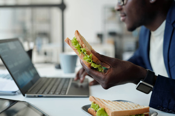 Focus sur sandwich végétarien appétissant en main du jeune manager afro-américain affamé en tenue de cérémonie à l'aide d'un ordinateur portable lors de la pause déjeuner - Photo, image