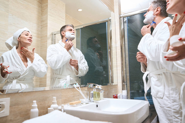 Couple in white coats standing near mirror, male shaves, female applies cream on skin in bathroom - Foto, imagen