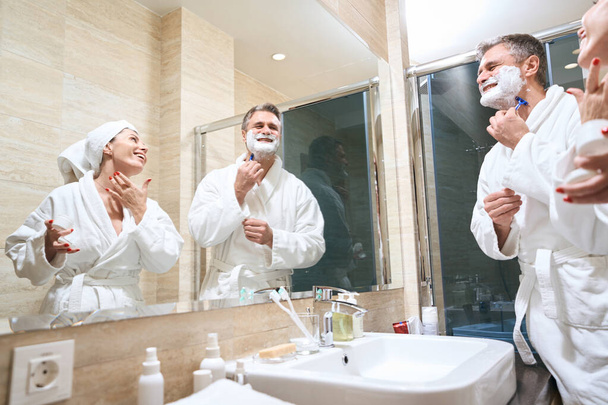 Husband and wife in white coats standing near mirror, man shaves, woman applies cream in the hotel - Photo, Image