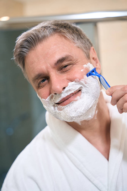 Smiling male in white coat shaving in the bathroom, looking at the camera - Photo, image