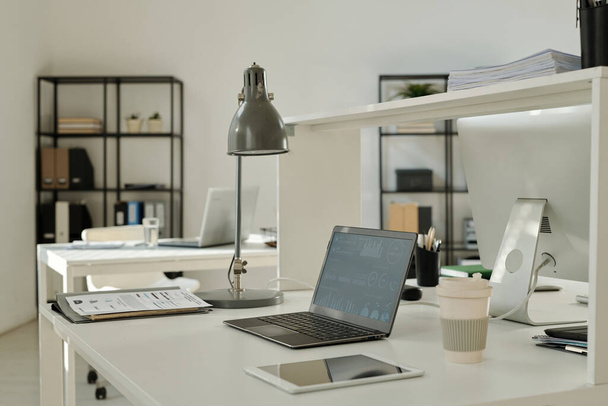 Финансовые графические данные на экране ноутбука, стоящего на столе среди бумажных документов, лампы, планшета и чашки кофе в просторном офисе - Фото, изображение