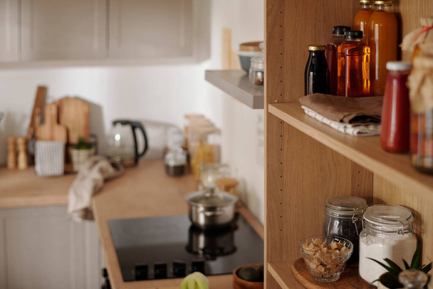 Деревянные полки с банками, содержащими муку и кофейные зерна, бутылки с огурцами, растительное масло, домашний сок и кетчуп на кухне - Фото, изображение