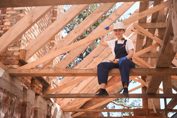 Cheerful high-qualified woman roofer sitting on wooden girder and looking at construction site, satisfied with done job and good materials - Foto, Bild