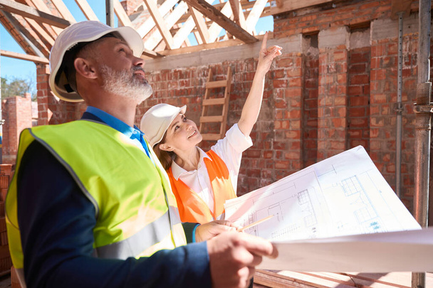 Woman architect showing somethimg on roof beams to serious foreman with blueprint, construction workers discussing building - Foto, Imagem