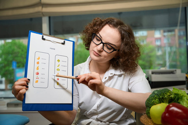 Curly-haired nutritionist demonstrates a visual aid with the nutritional characteristics of vegetables, next to a plate with colorful vegetables - Fotografie, Obrázek
