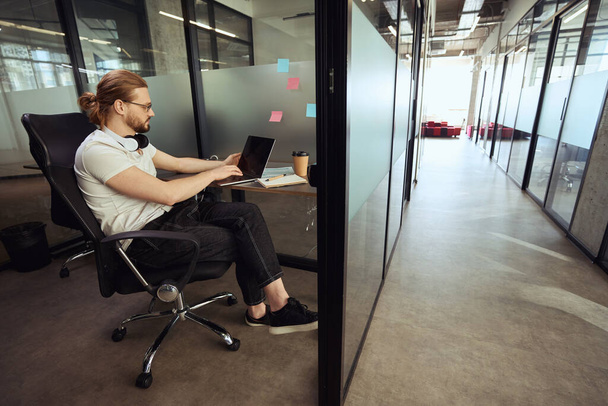 Boynunda kulaklığı olan genç bir çalışan rahat bir ofis bloğunda oturuyor ve bir dizüstü bilgisayarla çalışıyor. - Fotoğraf, Görsel