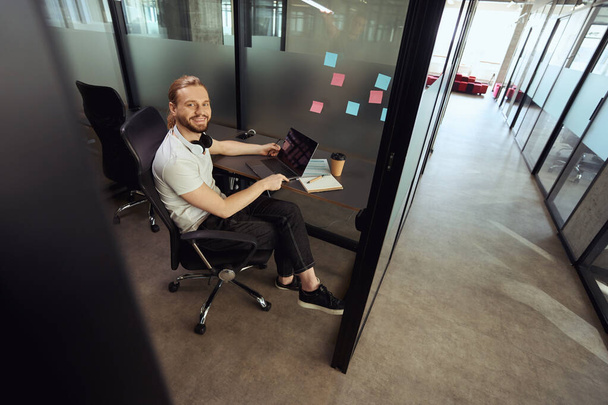 Handsome bearded guy comfortably settled down at his workplace in an office block, he works on a laptop - 写真・画像
