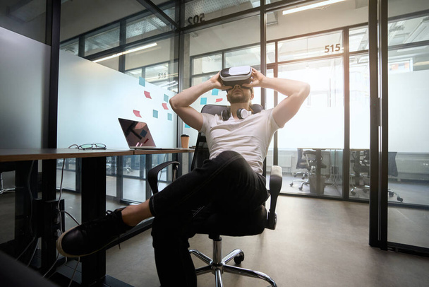 Bearded guy is sitting in a chair in an office box, he is using a virtual reality headset - 写真・画像