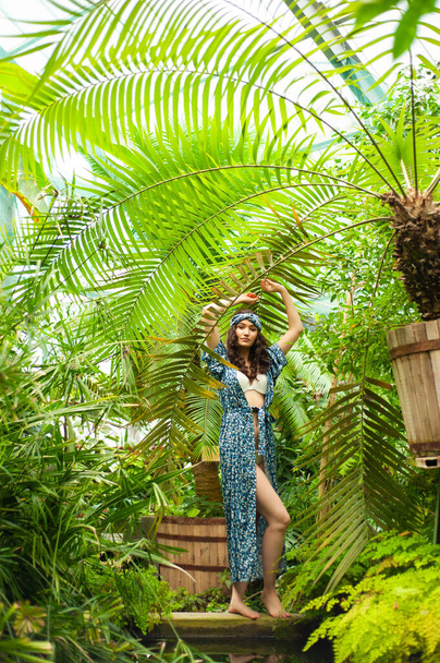 Gyönyörű lány zöld ruhában, kalapban és virágokkal. nő pózol egy trópusi dzsungelben. - Fotó, kép