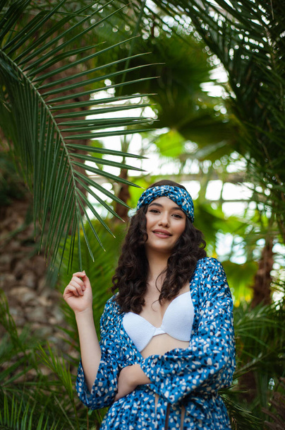 πορτρέτο μιας όμορφης γυναίκας με μπλε φόρεμα και καπέλο στο φόντο των δέντρων. τροπικά φύλλα. - Φωτογραφία, εικόνα