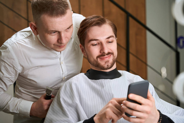 Male hairdresser and smiling man in cutting hair cape browsing cell phone in modern salon - Foto, Imagen
