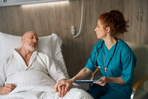 Doctora de uniforme con bloc de notas sentada y cogida de la mano de un paciente anciano en la cama en el hospital - Foto, imagen