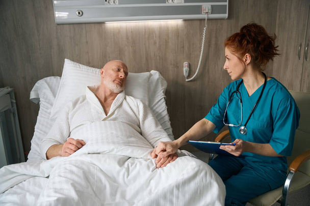 Beautiful medical practitioner with stethoscope encouraging her patient who lying on bed while holding his hand in clinic ward - Foto, imagen