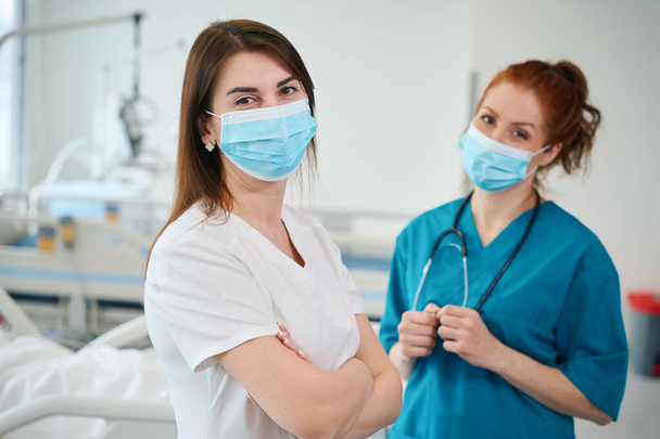 Retrato de duas belas médicas vestindo máscara médica e uniforme em pé na enfermaria do hospital - Foto, Imagem