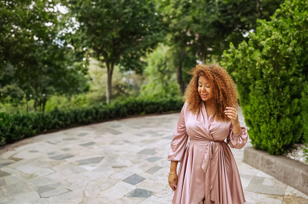 Молода красива афроамериканка насолоджується прогулянкою в літньому громадському зеленому парку. Щаслива жінка плюс розмір в шовковій рожевій сукні посміхається прогулюється по міських вулицях - Фото, зображення