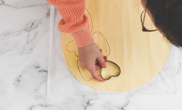 Hands of caucasian teenage girl cutting dough with metal curly heart cutter on white parchment with heart cutters on marble table, closeup flat lay. Cooking at home, homemade cookies and valentine's day concept. - Valokuva, kuva