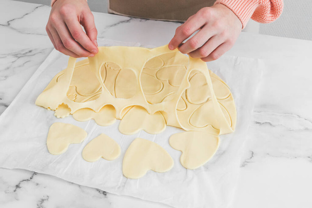 Hände einer jungen kaukasischen Bäckerin beim Trennen von rohem Teig mit Herzausschnitten auf weißem Pergament auf einem Marmortisch, Nahaufnahme von der Seite. Kochen zu Hause, hausgemachte Plätzchen und Valentinstag-Konzept. - Foto, Bild