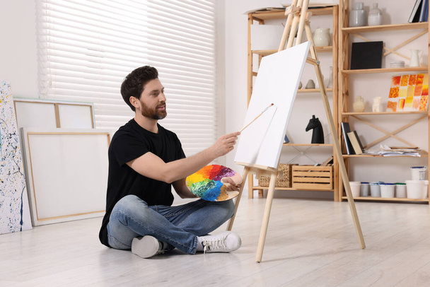 Man painting in studio. Using easel to hold canvas - 写真・画像