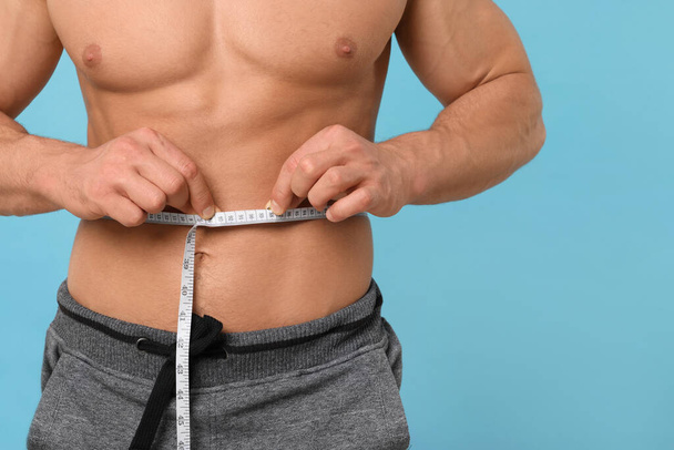 Athletic man measuring waist with tape on light blue background, closeup. Weight loss concept - Foto, afbeelding