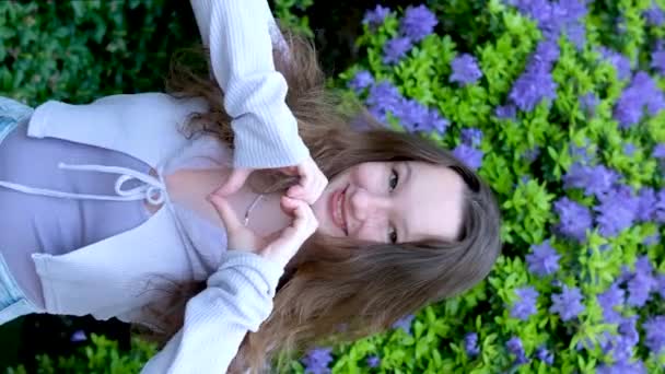 jong meisje toont een hart met haar handen lacht sterk schudt haar hoofd toont een duim oog. verticale video glimlach op de achtergrond van een blauwe struik van bloemen - Video