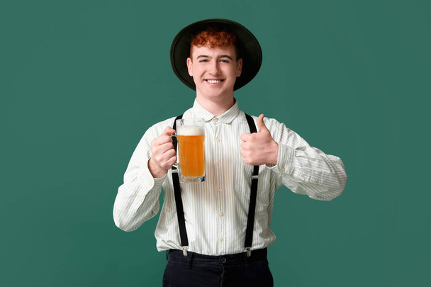 Nuori mies perinteisissä saksalaisissa vaatteissa, joissa olut näyttää peukaloa vihreällä taustalla - Valokuva, kuva
