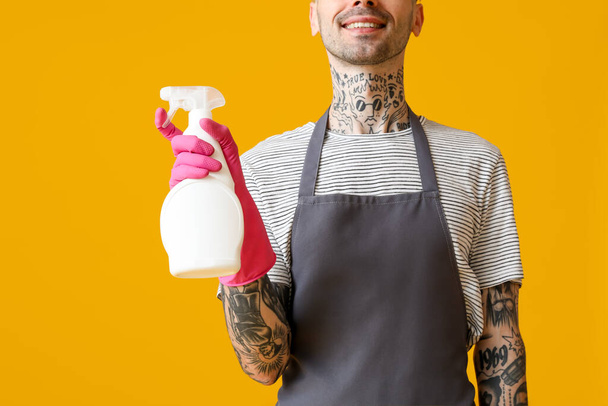 Young tattooed man with bottle of detergent on orange background, closeup - Photo, Image