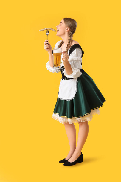 Hermosa camarera Octoberfest con cerveza comiendo salchicha sobre fondo amarillo - Foto, Imagen