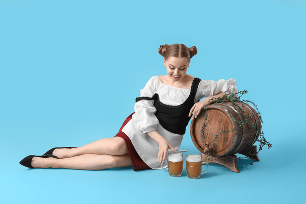 Belle serveuse Octoberfest avec bière et tonneau assis sur fond bleu - Photo, image