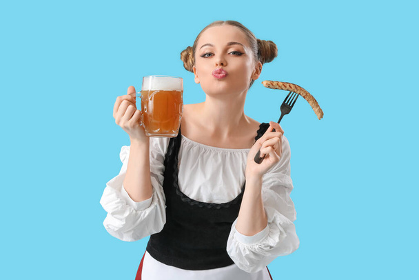 Όμορφη σερβιτόρα Octoberfest με μπύρα και λουκάνικο φυσώντας φιλί σε μπλε φόντο - Φωτογραφία, εικόνα