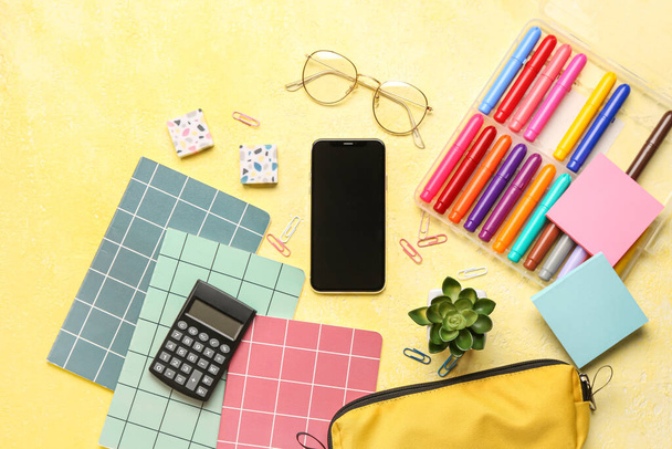 Мобильный телефон с очками и различными канцелярскими принадлежностями на гранж-желтом фоне - Фото, изображение