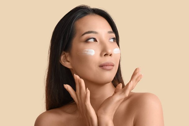 Belle jeune femme asiatique avec de la crème sur son visage sur fond beige - Photo, image