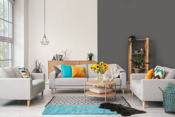 Gemütliche graue Sofas und Vase mit schönen Sonnenblumen im Inneren des hellen Wohnzimmers - Foto, Bild