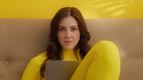Mediana toma de hermosa joven morena con el pelo largo bellamente peinado en traje amarillo sentado en el sofá, escribiendo en el ordenador portátil, lanzando miradas a la cámara. Imágenes de alta calidad 4k - Imágenes, Vídeo