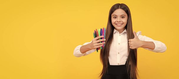 Happy girl dziecko trzymać filcowe końcówki długopisy pokazujące kciuki do góry gest żółty tło, zadowolony. Sztandar uczennicy. Portret ucznia szkoły z przestrzenią do kopiowania - Zdjęcie, obraz