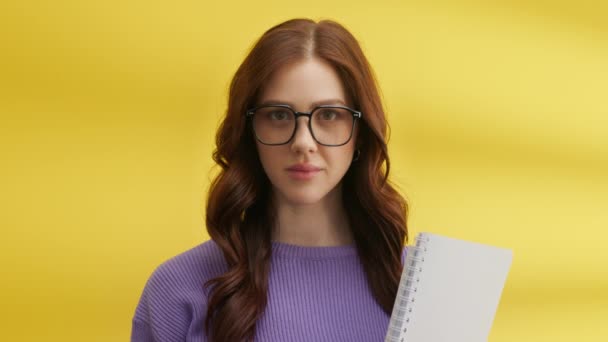 大きな眼鏡でかなり自信のある女性学生の中程度のショットは、春のノートを保持。黄色の背景に紫のニットセーターの若いかわいい女性。高品質4k映像 - 映像、動画