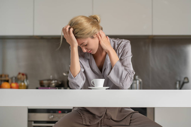 Расстроенная девушка в утренней пижаме с плохим настроением пьет кофе на кухне. Депрессивная женщина на кухне утром. Утро несчастной женщины. Грустная женщина - Фото, изображение
