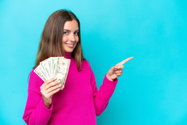 Young caucasian woman taking a lot of money isolated on blue background pointing finger to the side - Foto, imagen