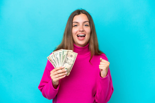 Young caucasian woman taking a lot of money isolated on blue background celebrating a victory in winner position - 写真・画像
