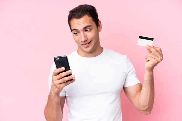 Молодий кавказький чоловік ізольований на рожевому тлі покупки з мобільним телефоном з кредитною карткою
 - Фото, зображення