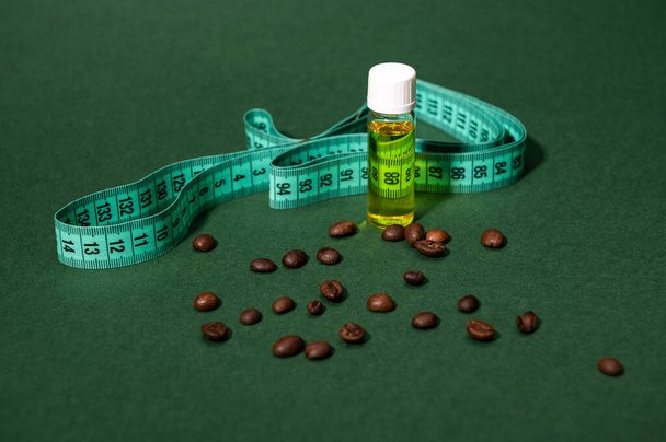 Still life with a transparent bottle of massage anti-cellulite oil, measure tape and coffee beans scattered on isolated green background. Slimming and weight loss concept. Cosmetology. Copy ad space - Photo, Image