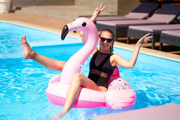 Hot slim woman have fun and wave hand on inflatable pink flamingo float mattress in bikini at swimming pool. Pretty female on tropical vacation. Attractive fit girl in swimwear lies in sun on floaty. - Photo, image