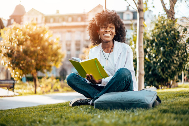 Beautiful and serious Black female student enjoying outdoors in college campus while study for the faculty exam. She is sitting on grass and reading something. Old building in background. - Φωτογραφία, εικόνα