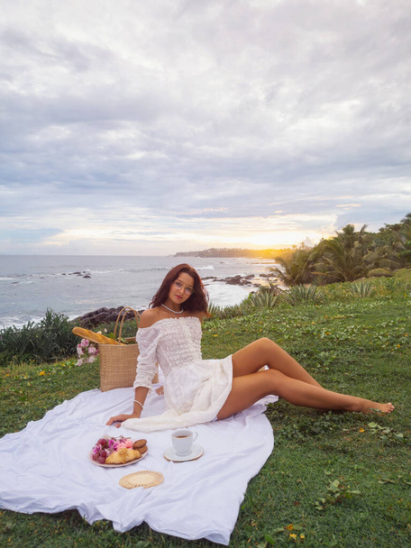 Picnics are good ideas for a romantic dating or creative photoshoot in boho style near the ocean on a green meadow. Chic woman in a white fashionable dress during a classical outdoor dinner with tea - Photo, image