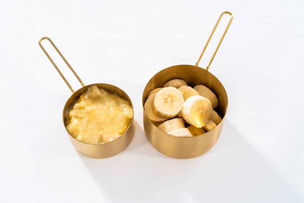 Mashed overripe bananas and sliced riped bananas in metal measuring cups. - Фото, изображение