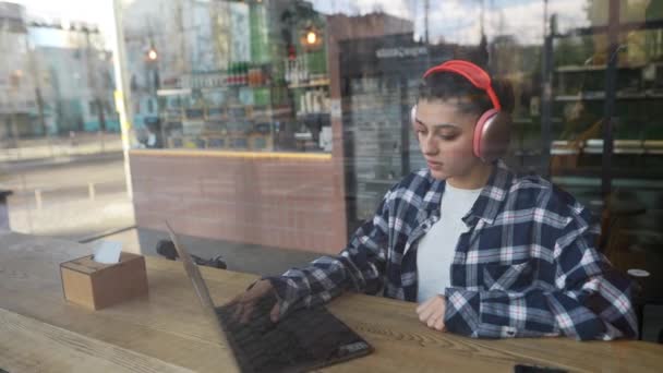 Gyönyörű lány fülhallgatóban gépel laptopon az ablak mögött a kávézóban. A kamera elé nézek. Kiváló minőségű FullHD felvételek - Felvétel, videó