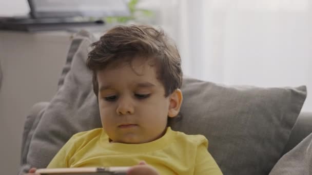Front view small kid boy enjoy using modern gadget smart phone looking at mobile screen, βλέποντας κινούμενα σχέδια, διασκεδάζοντας, παίζοντας κινητά online παιχνίδια, κάθονται στον καναπέ στο σπίτι. Κλείσε. - Πλάνα, βίντεο