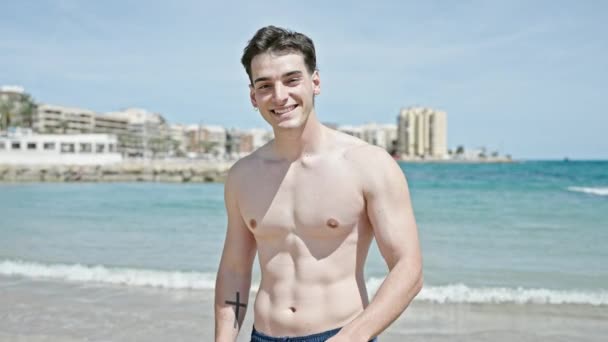 Fiatal spanyol férfi turista mosolygós magabiztos fésülködés haj kézzel a strandon - Felvétel, videó
