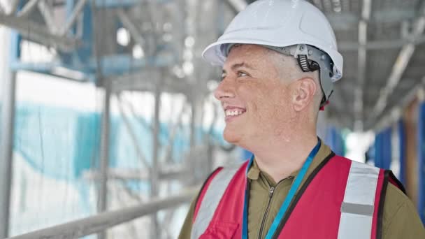 młody biały człowiek budowniczy uśmiech pewny siebie noszenie twardy kapelusz na miejscu budowy - Materiał filmowy, wideo