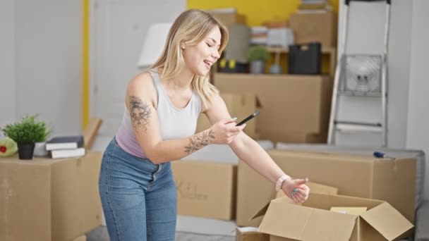 Νεαρή ξανθιά γυναίκα κάνει φωτογραφία σε πακέτα από το smartphone χαμογελώντας στο νέο σπίτι - Πλάνα, βίντεο