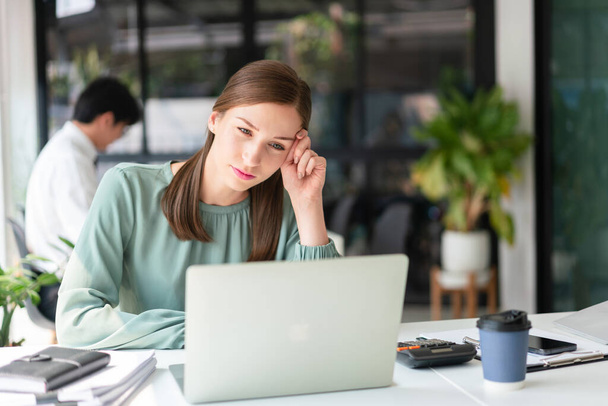 Femme d'affaires lisant des données financières sur ordinateur portable avec épuisé dans un lieu de travail moderne avec un collègue. - Photo, image